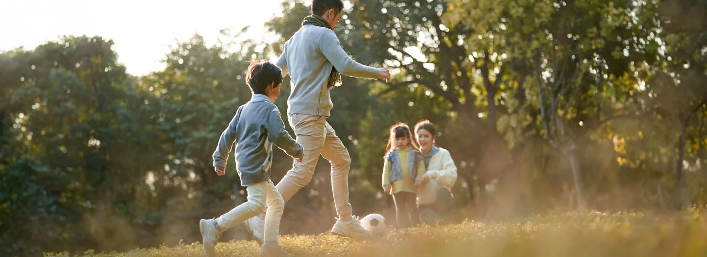 Father and son playing football in the park while the mother and daughter sit on the sidelines.