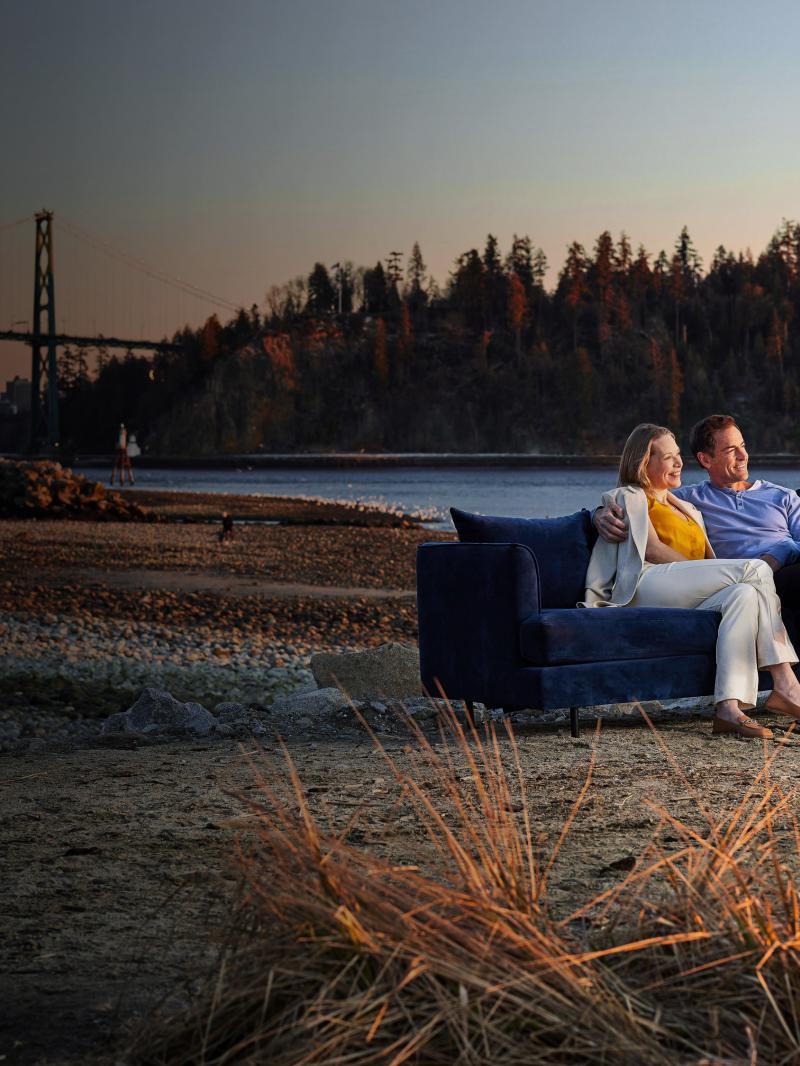 A couple sitting on a couch looking out at the water beside the Lions Gate bridge.