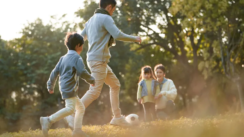 Father and son playing football in the park while the mother and daughter sit on the sidelines.